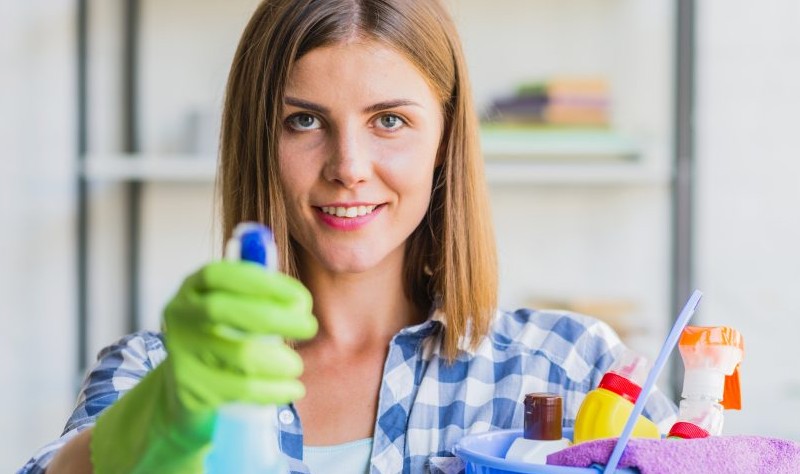 young woman holding a spray bottle and a bucket full of cloth rags and spray bottles