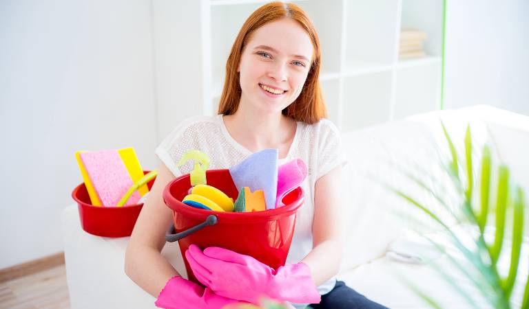 Woman in white top and pink gloves holding a red basket filled with tools and products