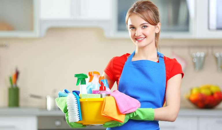 Woman in red-blue uniform and green gloves holding a yellow basket filled with bottles, tools, brushes.