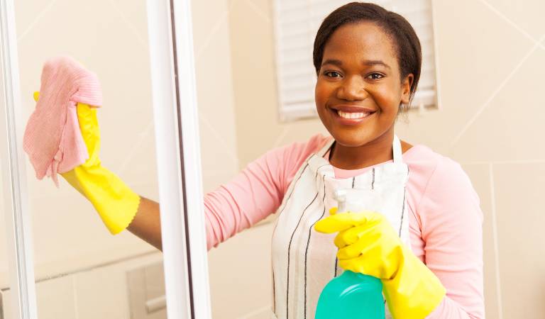 Woman in white-black dress and yellow gloves holding a green bottle cleaning shower door.