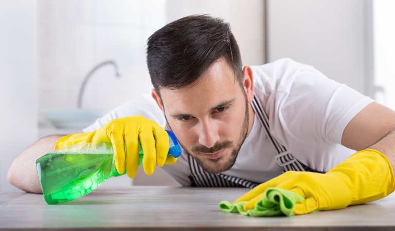 Man in white t-shirt and yellow glove holding a green bottle and cleaning surface with green cloth