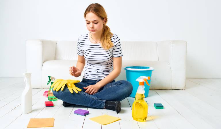 Woman is sitting on floor with cleaning bottles, gloves, cloths and blue basket.
