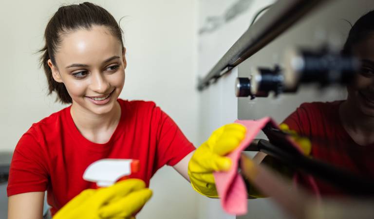 Woman in red top and yellow glove cleaning inside kitchen with a pink cloth.