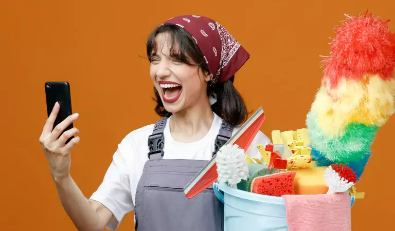 Woman looking on a phone and laughing and holding a bucket full of cleaning supplies