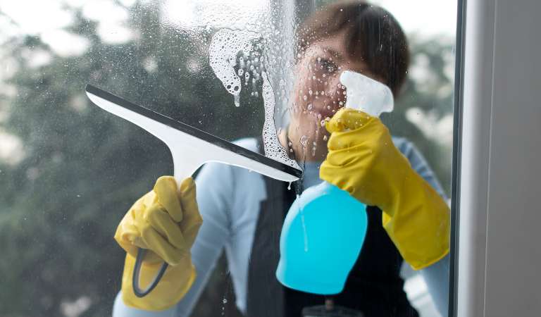 Woman in uniform and yellow gloves holding a blue spray bottle and cleaning outside window.