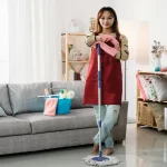 Cleaner standing smiling at camera while keeping her arms on the mop in bright home interior