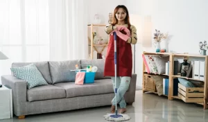 Cleaner standing smiling at camera while keeping her arms on the mop in bright home interior