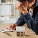 Stressed person holding a calculator with stacks of coins and a house model nearby