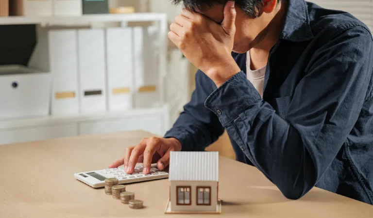 Stressed person holding a calculator with stacks of coins and a house model nearby